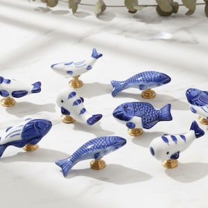 May include: A selection of ceramic fish-shaped cabinet knobs in blue and white. Each knob has a unique fish design, mounted on a gold-coloured base. The knobs are displayed on a white surface.