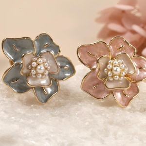 Könnte beinhalten: Nahaufnahme von zwei blütenförmigen Ohrringen. Ein Ohrring ist grau und weiß, der andere rosa und weiß. Beide haben einen goldenen Rand und eine Ansammlung kleiner Perlen in der Mitte. Die Ohrringe liegen auf einer weißen Oberfläche.