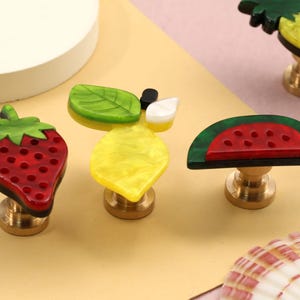 May include: A set of four fruit-shaped cabinet knobs featuring a red strawberry, a yellow lemon with green leaves, a red watermelon slice with a green rind, and a yellow pineapple, all mounted on circular brass-toned metal bases.