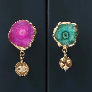 May include: Two decorative cabinet knobs featuring irregular, polished agate slices with gold-toned edges. One knob is vibrant magenta, the other is emerald green. Each knob has a gold-toned metal charm hanging from it: one an eye, the other stars.