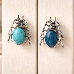 May include: Two beetle-shaped cabinet knobs attached to light-colored cabinet doors. The left knob features a bright turquoise oval stone with dark veining, while the right knob has a deep blue oval stone. Both have silver-toned metal legs.