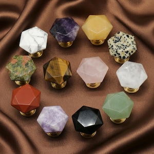 Könnte beinhalten: Eine Sammlung von Schrankknöpfen aus Edelsteinen in verschiedenen Farben und Mustern, darunter weißer Marmor, Amethyst, gelber Quarz und Rosenquarz. Jeder Knopf hat ein facettiertes Design und einen Messingfuß, ideal für Möbel.