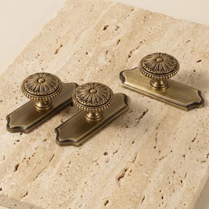 May include: Three antique brass cabinet pulls with a floral design on a rectangular backplate. The pulls have a vintage aesthetic and are likely for furniture restoration or home decor projects.