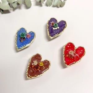 Puede incluir: Cuatro colgantes de resina en forma de corazón en azul, morado, rojo y marrón, cada uno con un borde dorado. Los colgantes están decorados con pequeñas flores secas y copos de oro, creando un aspecto único y artesanal.