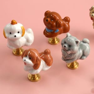 Puede incluir: Una colección de seis pomos de cerámica para cajones con forma de diversas razas de perros, cada uno con un acabado brillante y montado sobre una base metálica circular de tono dorado. Los perros aparecen en blanco, marrón y gris.