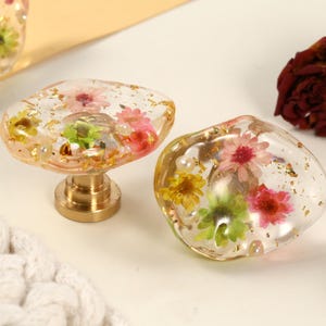 May include: Two clear resin cabinet knobs with embedded dried flowers in shades of pink, yellow, and green, and gold flakes. One knob is mounted on a brass base. Decorative hardware.