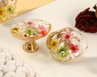 May include: Two clear resin cabinet knobs with embedded dried flowers in shades of pink, yellow, and green, and gold flakes. One knob is mounted on a brass base. Decorative hardware.