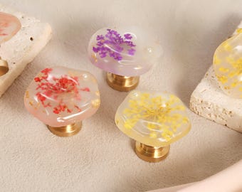 May include: Decorative cabinet knobs with brass bases and clear, organic-shaped tops. The knobs contain dried flowers in shades of red, purple, and yellow. Gold leaf accents are visible within the resin. These knobs are a unique home decor accent.