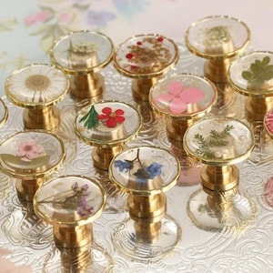 Op de afbeelding: Een verzameling decoratieve kastknoppen met transparante hars tops, elk met een andere gedroogde bloem of botanisch element. De knoppen hebben goudkleurige bases en zijn gerangschikt op een zilveren dienblad, met een verscheidenheid aan kleuren en texturen.