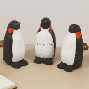 May include: Three penguin ornaments with black bodies, white bellies, and red details. Each penguin has a gold knob attached. The ornaments are displayed on a light-coloured surface.