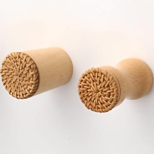 Op de afbeelding: Een reeks cilindrische wandhaken gemaakt van lichtgekleurd hout. Elke haak heeft een plat, cirkelvormig voorvlak ingelegd met een geweven rotanpatroon. De haken zijn horizontaal gemonteerd en hebben een gladde, natuurlijke houten afwerking.