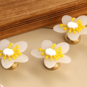 May include: A set of drawer knobs shaped like five-petaled flowers. Each knob features translucent white petals, a bright yellow stamen centre, and a white textured core, all mounted on a circular metallic gold base for attachment.