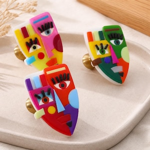 May include: Three decorative cabinet knobs featuring abstract face designs in vibrant multi-colored acrylic. Each knob has a brass-toned metal base and displays geometric shapes, bold lines, and stylized eyes in yellow, pink, green, red, and blue.