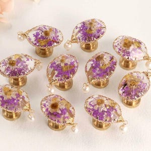 May include: A collection of nine drawer knobs featuring clear resin oval tops embedded with dried purple and white flowers. Each knob has a gold-toned metal base and is accented with a small white pearl bead attached by gold wire to the side.