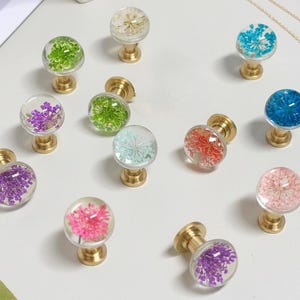 May include: A collection of decorative cabinet knobs with clear, spherical resin tops. Each knob features a different dried flower arrangement in shades of purple, pink, green, blue, and orange. The knobs have brass-colored bases.