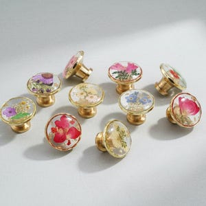 May include: A selection of decorative cabinet knobs with floral designs. Each knob has a clear, rounded top encasing dried flowers in various colours, set on a gold-toned base. The knobs are arranged on a light-coloured surface.