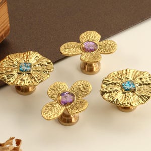 Peut inclure: Quatre boutons de meuble décoratifs avec un motif floral. Les boutons sont de couleur dorée avec une finition texturée. Deux boutons ont un centre carré de couleur turquoise et deux ont un centre violet. Les boutons sont en métal.
