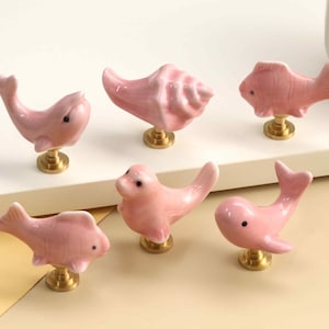 May include: A set of six pink ceramic cabinet knobs featuring various sea-themed shapes including fish, a seashell, and a seal. Each knob is attached to a small, circular, metallic gold-toned base, providing a consistent look for furniture hardware.