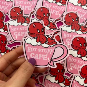 Puede incluir: Una taza de café rosa con un bebé diablo rojo sentado en el borde. La taza tiene espuma blanca y el texto "Hot Stuff Cafe" en letras rosas.
