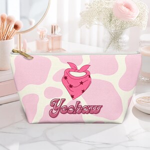 Op de afbeelding: Een etui met rits voorzien van een lichtroze en crèmekleurig koeienvlekkenpatroon. In het midden staat een afbeelding van een felroze bandana met kleine donkere sterren en het woord "Yeehaw" in een cursief, donkerroze lettertype.