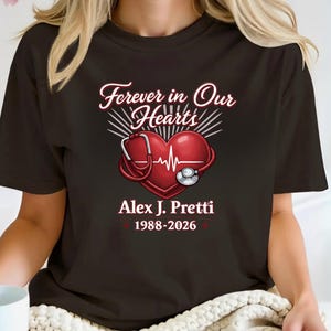 May include: Black t-shirt featuring a red heart with a stethoscope and an EKG line. The text reads "Forever in Our Hearts," the name "Alex J. Pretti," and the years "1988-2026."