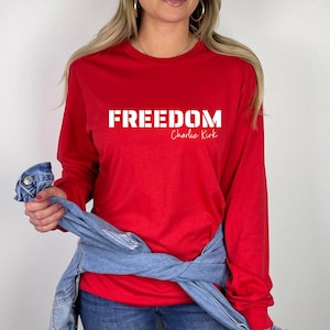 Puede incluir: Una camiseta roja de manga larga con la palabra "FREEDOM" impresa en letras blancas. El nombre "Charlie Kirk" está impreso debajo en cursiva. Una chaqueta vaquera está sobre los hombros de la persona.