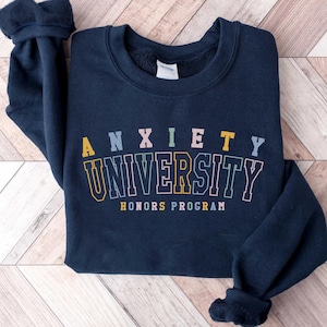 Könnte beinhalten: Marineblaues Sweatshirt mit dem Schriftzug "ANXIETY UNIVERSITY HONORS PROGRAM" in bunten Blockbuchstaben. Das Sweatshirt ist gefaltet, die Ärmel sind sichtbar. Der Text ist in Gelb, Rosa, Grün und Blau.