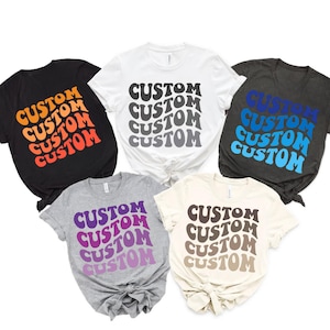 May include: Five t-shirts with the word "CUSTOM" repeated in a wavy, retro font. The shirts are in different colors: black, white, gray, heather gray, and beige. The text on the shirts is in different colors, creating a gradient effect.