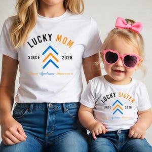 May include: Two white t-shirts with matching designs. One shirt says "LUCKY MOM" with a blue and orange arrow design and "Down Syndrome Awareness." The other says "LUCKY SISTER" with the same design.