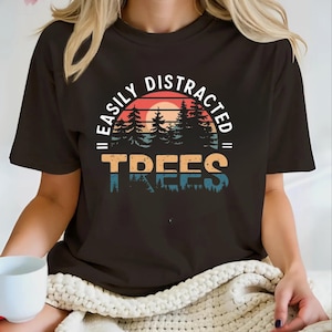 May include: Black t-shirt with the text "EASILY DISTRACTED TREES" in a retro design. The graphic features a sunset, trees, and a color palette of orange, blue, and green.