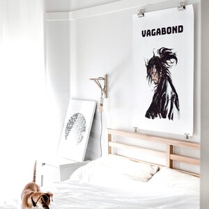 May include: A white bedroom with a bed, a white nightstand, and a white lamp. A black and white print of a circle is hanging on the wall. A poster with the word "VAGABOND" in black text is hanging on the wall above the bed. A brown and white dog is standing on the floor in front of the bed.