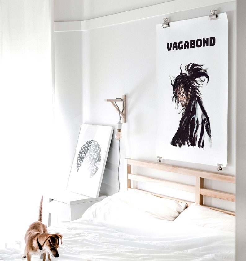 May include: A white bedroom with a bed, a white nightstand, and a white lamp. A black and white print of a circle is hanging on the wall. A poster with the word "VAGABOND" in black text is hanging on the wall above the bed. A brown and white dog is standing on the floor in front of the bed.