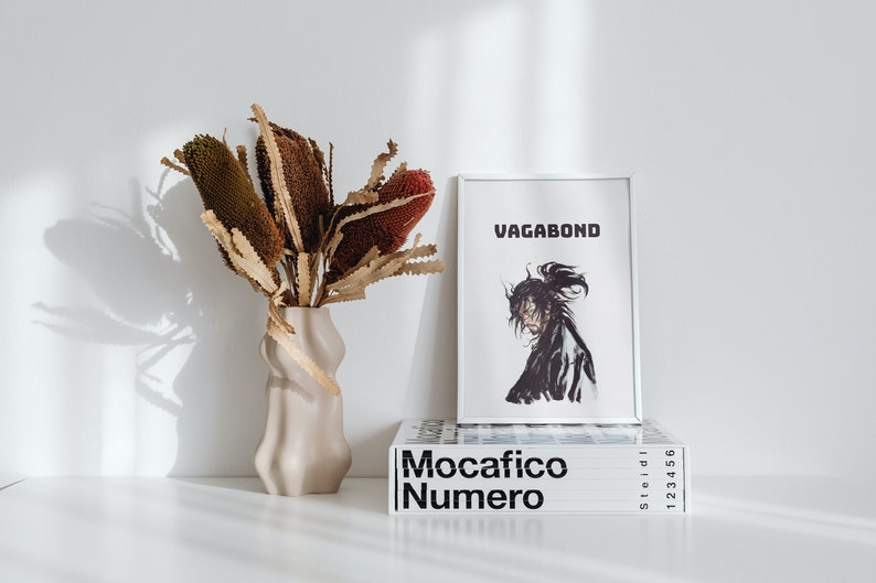 May include: A white framed print with the word "Vagabond" in black text. The print features a black and white illustration of a person with long hair. The print is sitting on top of a stack of books with the title "Mocacifico Numero" in black text.