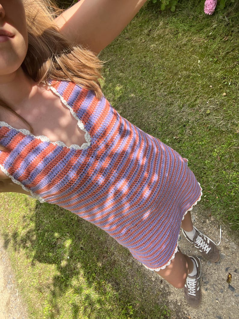 May include: A handmade crochet dress featuring alternating stripes of orange and lavender. The dress has a halter neckline and a scalloped edge. The dress is worn with brown sneakers.