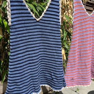 May include: Two handmade crochet tops hanging on a clothesline. One is blue with navy stripes, the other is red with purple stripes. Both have a scalloped edge and a V-neck design. The tops are made of yarn.