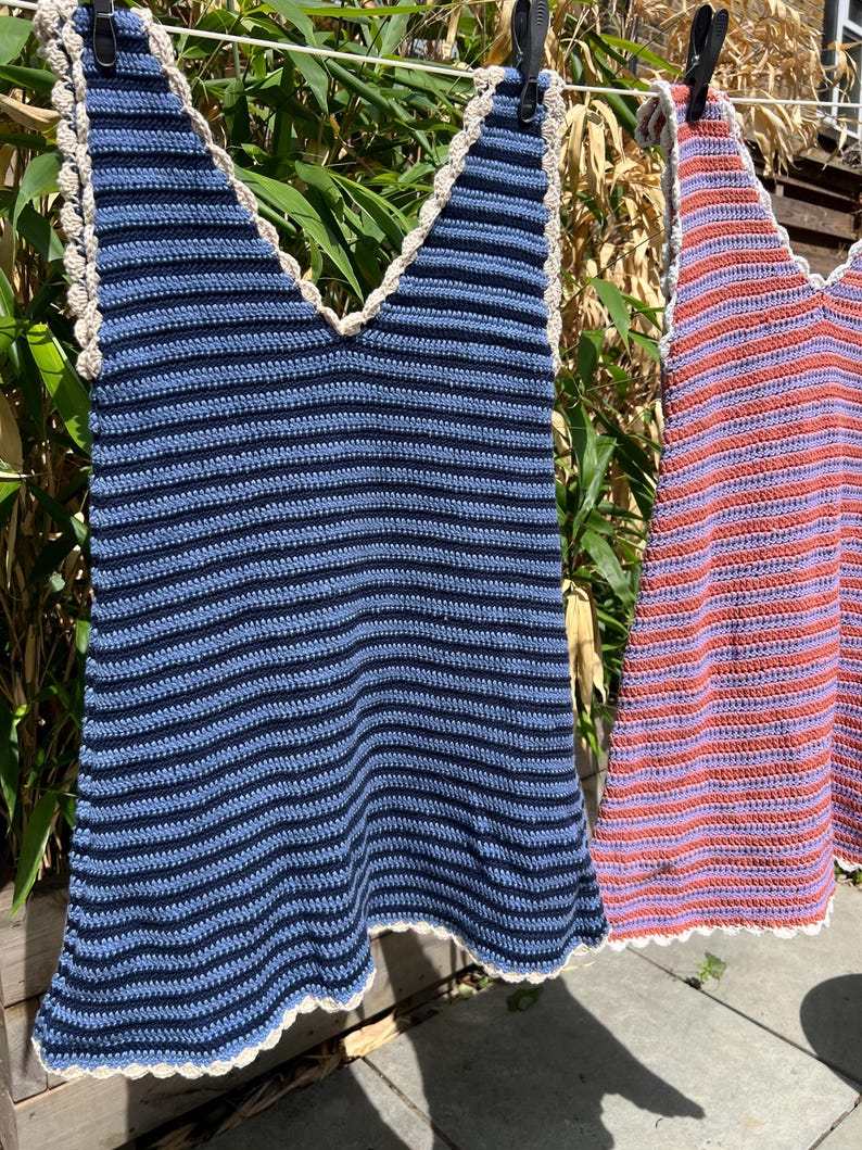 May include: Two handmade crochet tops hanging on a clothesline. One is blue with navy stripes, the other is red with purple stripes. Both have a scalloped edge and a V-neck design. The tops are made of yarn.