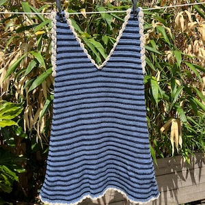 May include: A blue and navy striped crochet top with a V-neck design. The top has a cream-colored crocheted trim along the neckline and hem. It is hanging on a clothesline in front of green foliage.