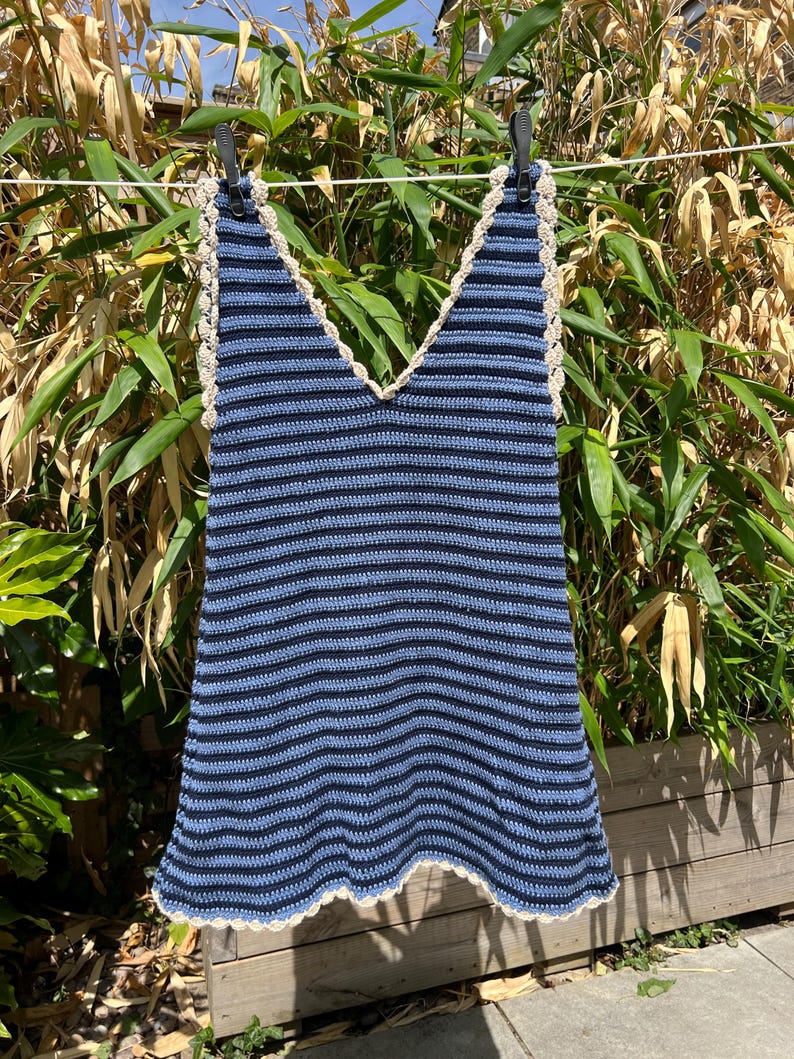 May include: A blue and navy striped crochet top with a V-neck design. The top has a cream-colored crocheted trim along the neckline and hem. It is hanging on a clothesline in front of green foliage.