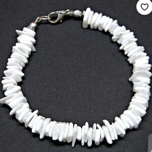 Puede incluir: Pulsera de chips blancos con cierre plateado. La pulsera está hecha de pequeños chips blancos de forma irregular unidos entre sí. El cierre es de tipo mosquetón estándar. Fondo oscuro.