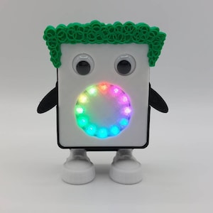 Könnte beinhalten: Ein weißes Roboter-Spielzeug mit einer grünen Haarpracht, schwarzen Armen und weißen Schuhen. Der Roboter hat große schwarze Augen und ein kreisförmiges Regenbogen-LED-Licht in der Mitte seines Gesichts.