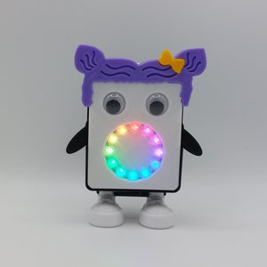 Könnte beinhalten: Ein weißer Spielzeugroboter mit einer lila Perücke, einer gelben Schleife, schwarzen Armen und weißen Füßen. Der Roboter hat große schwarze Augen und ein Regenbogen-LED-Licht in der Mitte seines Gesichts.