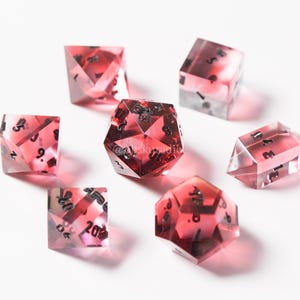 May include: Set of seven red and clear dice with black numbers. The dice are made of resin and have a variety of shapes, including a d4, d6, d8, d10, d12, and d20.