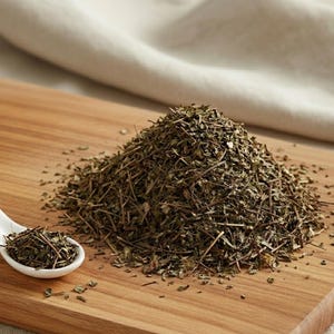 Può includere: Un tagliere in legno con un mucchio di erbe essiccate e un cucchiaio di ceramica bianca pieno di erbe. Le erbe sono un mix di verde e marrone, e il cucchiaio è a sinistra del mucchio. Un panno bianco sullo sfondo.