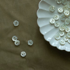 May include: A white ceramic plate with a scalloped edge holds a collection of white pearl-like buttons. The buttons are arranged in a random pattern and have a flower-like design.  There are also several buttons scattered on a brown surface.
