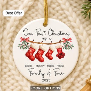 May include: A white ceramic Christmas ornament with a gold string for hanging. The ornament features the text "Our First Christmas as a Family of Four 2025." Below the text are four red stockings, each with a name. The words "Best Offer" are at the top.