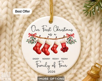 May include: A round white ceramic ornament with a gold hanging string. The design features four red Christmas stockings hanging from a branch with mistletoe. Text reads Our First Christmas as a Family of Four, names Daddy, Mommy, Rocky, Roony, and 2026.
