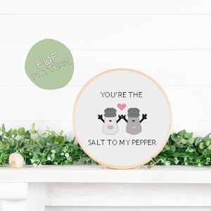 Puede incluir: Un aro de bordado blanco con un diseño de punto de cruz que presenta dos saleros y pimenteros tomados de la mano con un corazón entre ellos. El texto "You're the salt to my pepper" está bordado debajo de los saleros.