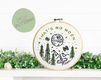May include: Embroidered hoop art with a white background and the text "That's no moon" in green lettering. The design features a stylized Death Star from Star Wars, surrounded by green pine trees and a white background with gold stars.