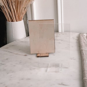 May include: A clear acrylic picture frame with a wooden backing on a white marble surface. A bouquet of dried pampas grass is in the background.