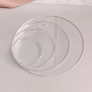 May include: A set of three clear acrylic circles of varying sizes, nested together. The largest circle has a small hole near the edge. The circles are transparent and appear to be made of plastic. The objects are on a light gray surface.